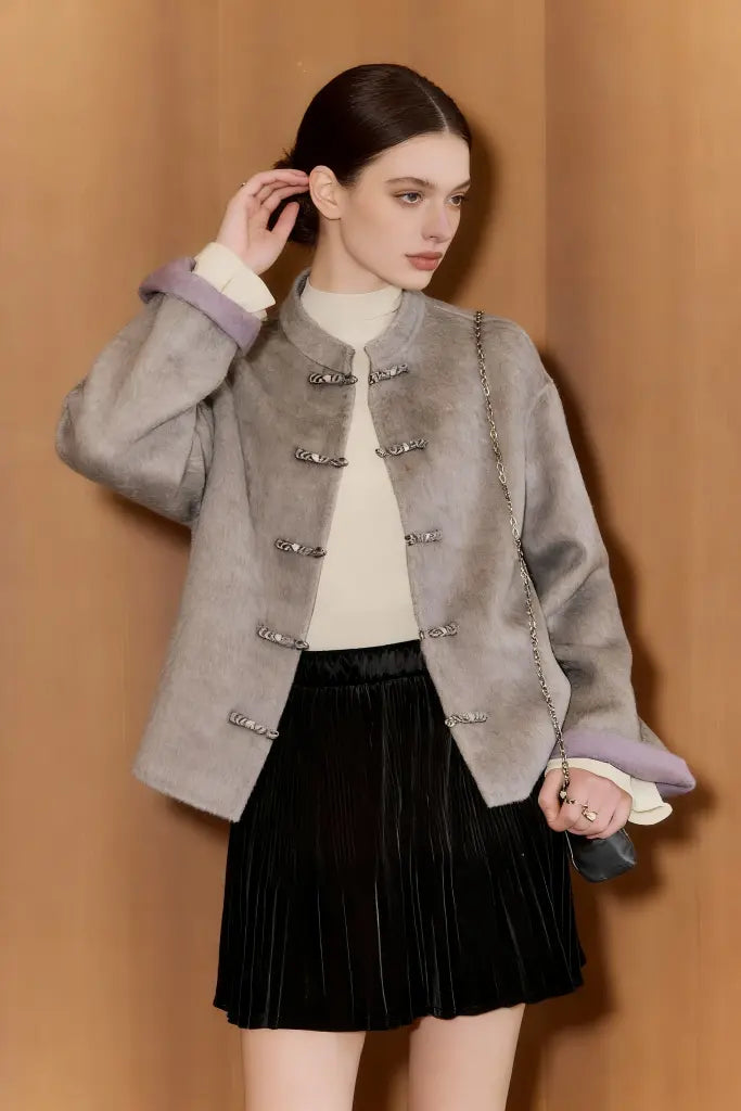 Woman wearing a gray textured jacket with a beige sweater and black pleated skirt against a beige background