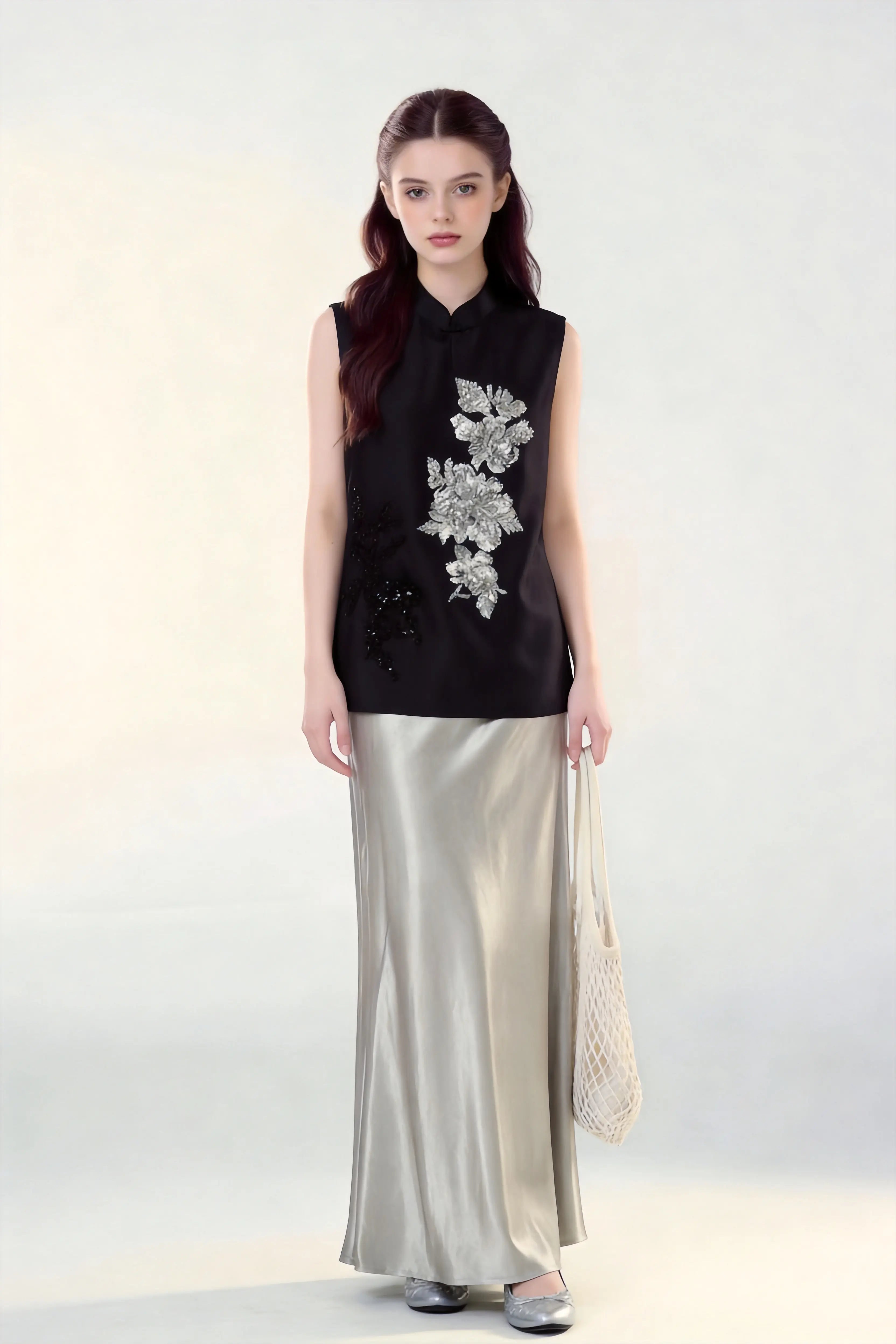 Woman wearing a black Beaded Lotus Embroidery Loose Sleeveless Top & Satin Skirt Two-Piece Set, holding a woven bag on a white background.