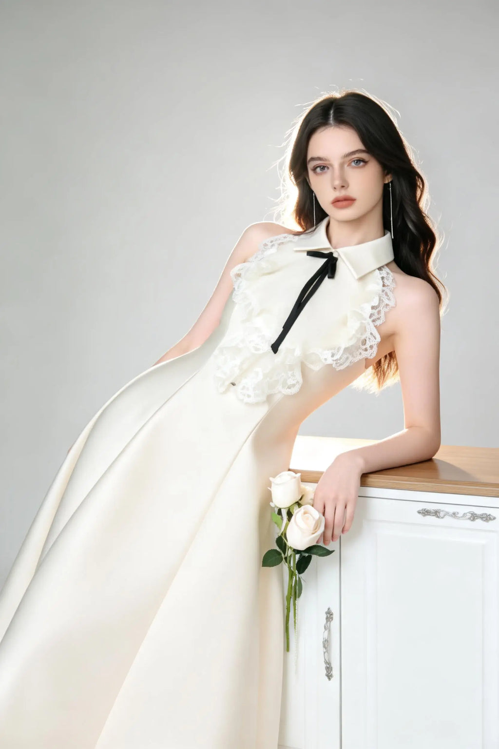 Woman in a white elegant halter neck satin flared a-line dress with lace collar and bow detail standing against a gray background