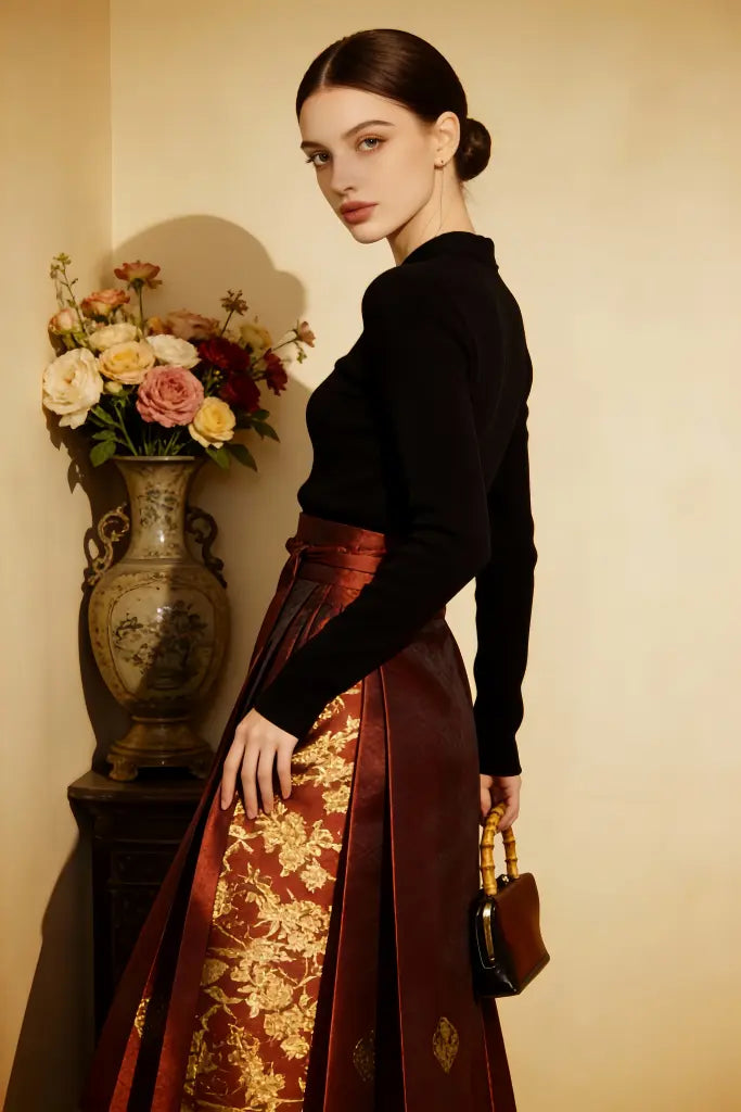 Woman in a black top and red floral skirt standing next to a vase with flowers on a beige wall.